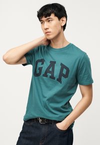 Νέος άνδρας με τιρκουάζ μπλουζάκι GAP, μαύρη ζώνη και σκούρο τζιν, στηρίζοντας το ένα χέρι στον λαιμό και το άλλο στην τσέπη, μπροστά σε απλό φόντο.