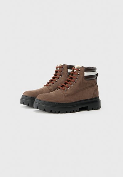 Bottines en suède marron avec des semelles épaisses noires à crampons, des lacets rouges et des cols rembourrés avec des rayures blanches et marron foncé.