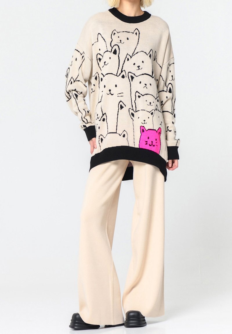 Personne portant un pull beige oversize avec des poignets et un col noirs, orné de plusieurs visages de chats esquissés en noir et d'un chat rose vif, associé à un pantalon large beige et des chaussures noires.