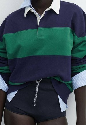 Osoba ma na sobie granatowo-zieloną, pasiastą koszulkę rugby na niebiesko-białej, pasiastej koszuli z kołnierzykiem, w połączeniu z czarnymi sportowymi szortami ze ściągaczem.