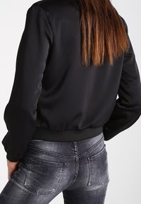 Black satin bomber jacket with a fitted waist, long sleeves, and ribbed cuffs. Paired with gray distressed skinny jeans.