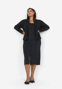 Cardigan nero con sottili brillanti sopra una maglietta nera e una gonna a tubino nera fino al ginocchio con uno spacco frontal. Accentuato da tacchi neri a fiocco.