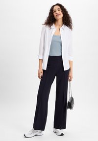 Witte button-up shirt over een lichtblauwe tanktop, gecombineerd met donkere wijde broek en witte sneakers; met een kleine zwarte handtas.