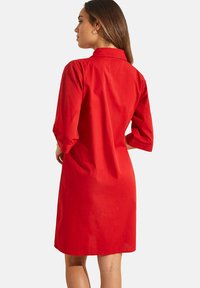 Robe en coton rouge avec un col et des manches trois-quarts, présentant une coupe droite et une texture lisse. Design simple sans motifs visibles.