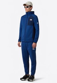 Niebieska kurtka zapinana na suwak z ciemnymi akcentami, dopasowane spodnie dresowe i detale z logo. Akcesoria to jasna czapka i okulary przeciwsłoneczne. Lekki materiał.