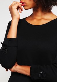Maglione lavorato a maglia nera con scollo rotondo e polsini risvoltati, rifinito con dettagli in metallo argentato. Tessuto morbido e design aderente.