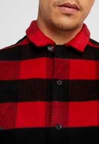 Röd och svart rutigt flanellskjorta med en mjuk, texturerad krage; har mörka knappar och en lagerdesign i botten.