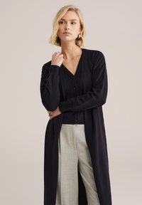 Cardigan nero con scollo a v, maniche lunghe e bottoni, indossato sopra una maglia nera, abbinato a pantaloni larghi di colore chiaro.