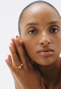 Anillo de oro con tres acentos esféricos, textura suave. El sujeto apoya una mano en su cara, destacando el diseño del anillo.