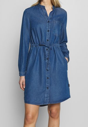 Vrouw in een knielange blauwe denim jurk met lange mouwen, voorknopjes, borstzak en een gestrikt tailleband.