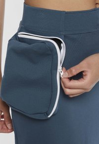 Pantalon de sport bleu sarcelle avec une poche zippée blanche. La poche est légèrement rembourrée et se ferme avec une fermeture éclair. Le tissu semble extensible et lisse.