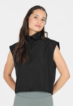Femme aux longs cheveux foncés portant un haut noir sans manches zippé et un pantalon ajusté gris clair, debout devant un fond uni.