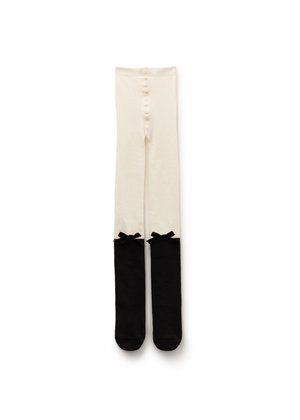 Collants pour enfants de couleur crème avec pieds noirs et petits nœuds noirs au sommet de la partie noire, étalés à plat sur un fond blanc.
