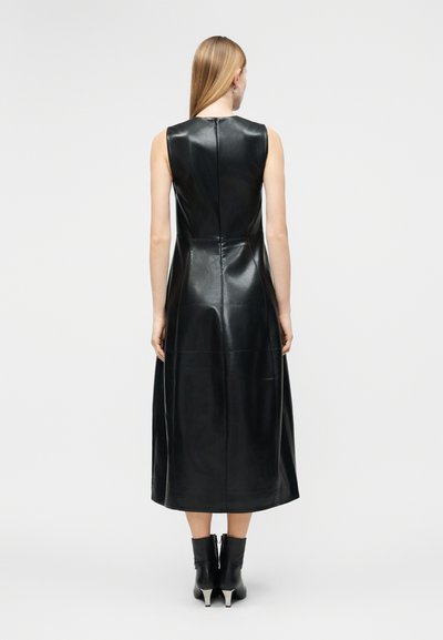 Γυναίκα με μακριά μαλλιά, γυρισμένη προς τα πίσω, φοράει μαύρο δερμάτινο μίντι φόρεμα χωρίς μανίκια και μαύρα μποτάκια με τακούνι σε λευκό φόντο.