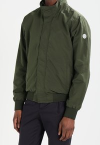 Groene bomberjack met een hoge kraag, ribgebreide manchetten en zoom. Gemaakt van lichtgewicht materiaal, met een logopatch op de linker mouw.