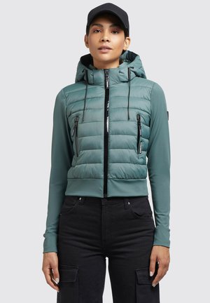 Veste matelassée courte en teal avec capuche, zip sur le devant et deux poches poitrine. Manches longues avec une finition en tissu lisse. Accents noirs.