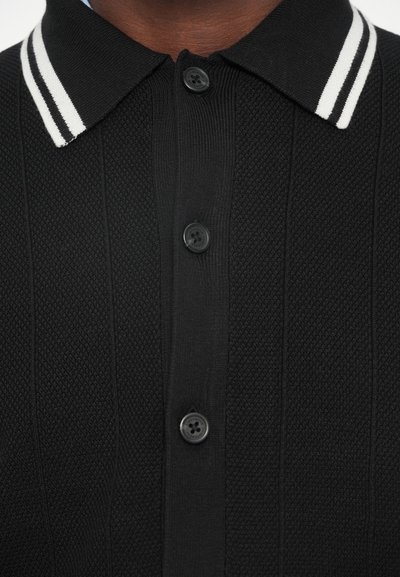 Musta poolpaita, jossa on teksturoitu kangas, valkoinen raidallinen kaulus ja neljä mustaa nappia napitettuna.