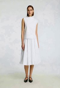 Weißes ärmelloses Baumwolloberteil kombiniert mit einem fließenden weißen Rock, beide mit minimalistischem Design und glatter Textur. Schwarze Ballerinas vervollständigen das Outfit.