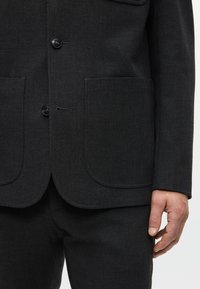 Giacca da completo grigio scuro con due grandi tasche a toppa, chiusura a tre bottoni e tessuto leggermente texturizzato. Design slim fit con linee pulite.