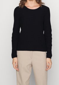 Maglione lungo a maniche, nero e a coste, con scollatura rotonda; abbinato a pantaloni beige. La texture è liscia e aderente, con un design minimale.