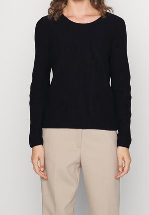 Pull noir à manches longues côtelé avec un col rond ; associé à un pantalon beige. La texture est lisse et ajustée, avec un design minimal.