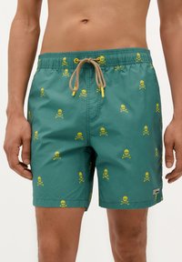 Shorts de bain verts en tissu léger, arborant un motif répété de têtes de mort et de tibias jaunes, avec une taille élastique à cordon.