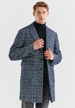 Navyi i białą kratkę wełniany płaszcz z wyłogowym kołnierzem, jednorzędowy fason i dużymi guzikami. Model ma na sobie czarny golf pod spodem.