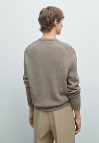 Brązowy sweter z dzianiny o fakturowanym wykończeniu, z prążkowanymi mankietami i dołem, z okrągłym dekoltem i rękawami raglanowymi. Nosi się go na beżowych spodniach.