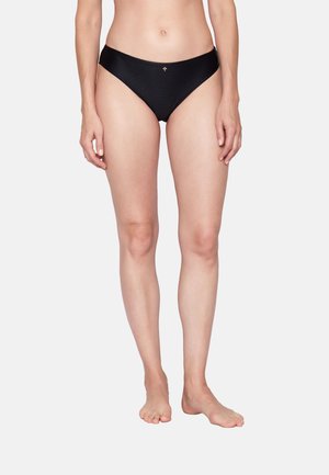 Frau trägt schwarze, gerippte Bikinihöschen mit kleinem goldenem Emblem, steht barfuß vor schlichtem hellem Hintergrund.