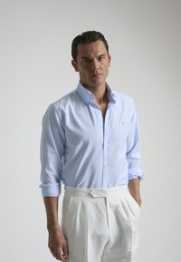 Camisa de botones azul claro con cuello, mangas largas enrolladas en los puños y una textura sutil, combinada con pantalones blancos con pliegues.