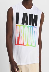 T-shirt blanc sans manches avec un grand texte noir "I AM" et un texte "PROUD" en dégradé arc-en-ciel. Tissu doux, coupe décontractée, détails minimalistes.