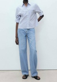 Chemise à boutons avec rayures bleues et blanches, manches retroussées ; associée à un jean large taille haute bleu clair ; sandales plates noires.