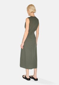 Ärmelloses olivgrünes Midikleid mit schmaler Taille und fließendem Stoff. Getragen mit schwarzen Rutschen-Sandalen. Die Rückansicht hebt die klaren Linien hervor.