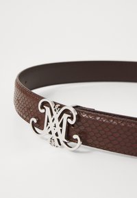 Cintura in pelle marrone testurizzata con grande fibbia a logo "M" intrecciata in argento su uno sfondo bianco semplice.