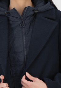 Námořnický kabát s kapucí, vybavený zipem a druky. Materiál je měkký a strukturovaný s vrstveným designem. Vnitřek je prošitý.