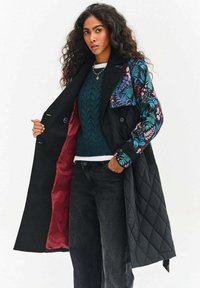 Dlhý čierny kabát s rukávmi s kvetinovou potlačou, prešívanou textúrou a vínovo červenou podšívkou, nosený cez tmavozelený pletený sveter a čierne džínsy.