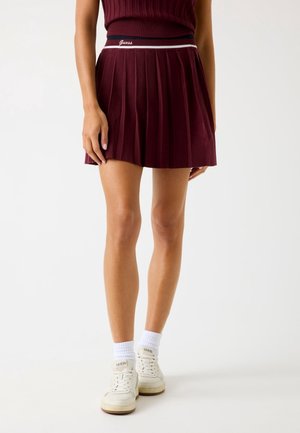 Mannequin portant une mini-jupe plissée bordeaux avec une ceinture à logo Guess, associée à des baskets blanches Guess et des chaussettes blanches aux chevilles.