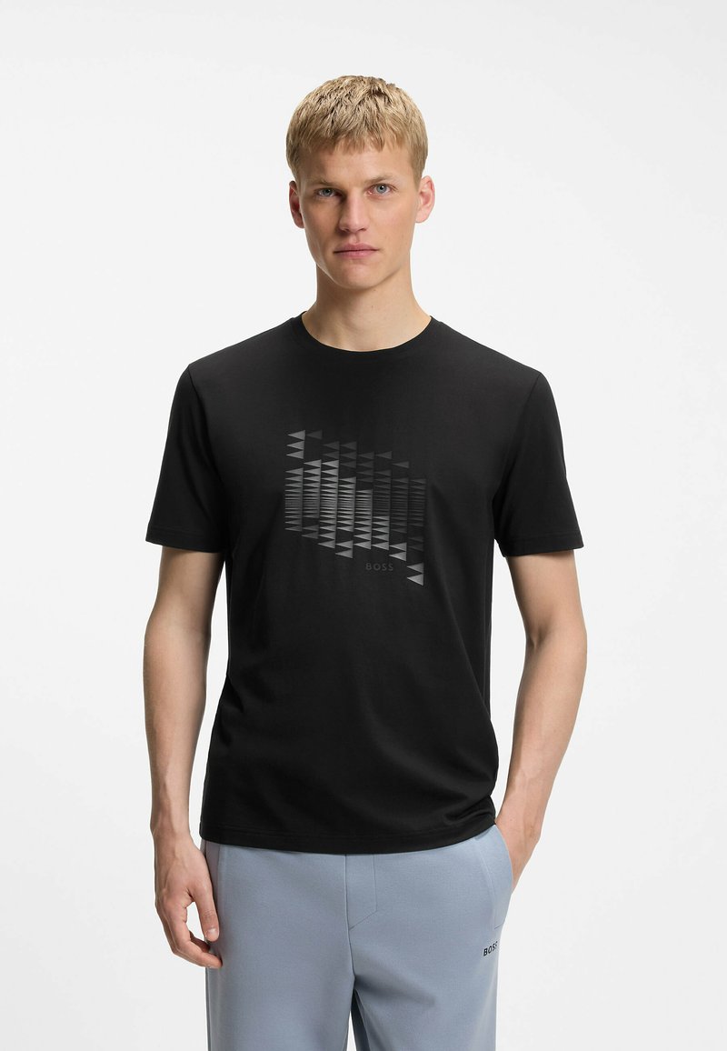 Homme portant un t-shirt noir à motif géométrique et un pantalon gris clair, debout devant un fond blanc uni.