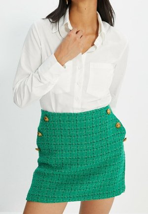 Grüner, strukturierter Minirock mit goldenen Knopfdetails, kombiniert mit einem weißen Button-up-Shirt. Der Rock hat ein gewebtes Muster und seitliche Knöpfe.