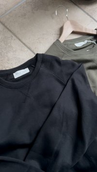 Two folded crewneck sweatshirts, one black labeled "Pier One" and one olive green on a wooden hanger on a tiled floor.