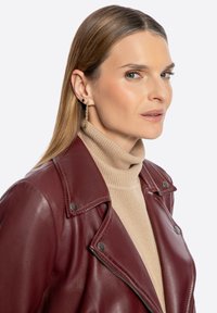 Giacca in pelle bordeaux con dettagli in argento, indossata sopra un dolcevita beige. Capelli lunghi e lisci, con orecchini pendenti d'oro.