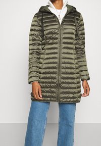 Persoon in een olijfgroene gebate lange hoodie-jack met zwarte koordjes en blauwe jeans, tegen een effen lichte achtergrond.