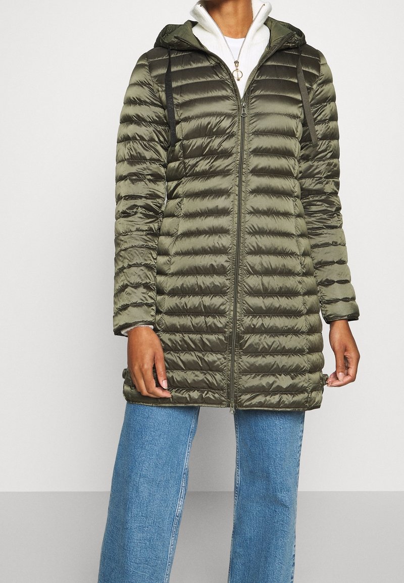 Persoon in een olijfgroene gebate lange hoodie-jack met zwarte koordjes en blauwe jeans, tegen een effen lichte achtergrond.