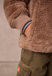 Persona che indossa una giacca marrone pelosa con la mano in tasca, sovrapposta a una manica blu, e pantaloni cargo verde oliva con una toppa arancione.