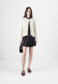 Kremfarget strikket cardigan med rund hals, treknapper, kombinert med et svart plissert skjørt; modellen holder en liten svart håndveske.