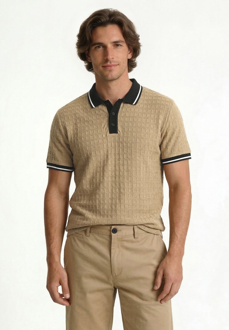 Homme aux cheveux mi-longs portant un polo beige texturé à manches courtes avec des bordures noires et un pantalon beige, debout sur fond blanc.