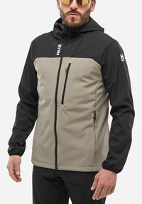Veste à capuche noire et beige, en matériau softshell. Elle possède une poche poitrine zippée, des poignets réglables et un ourlet élastique.