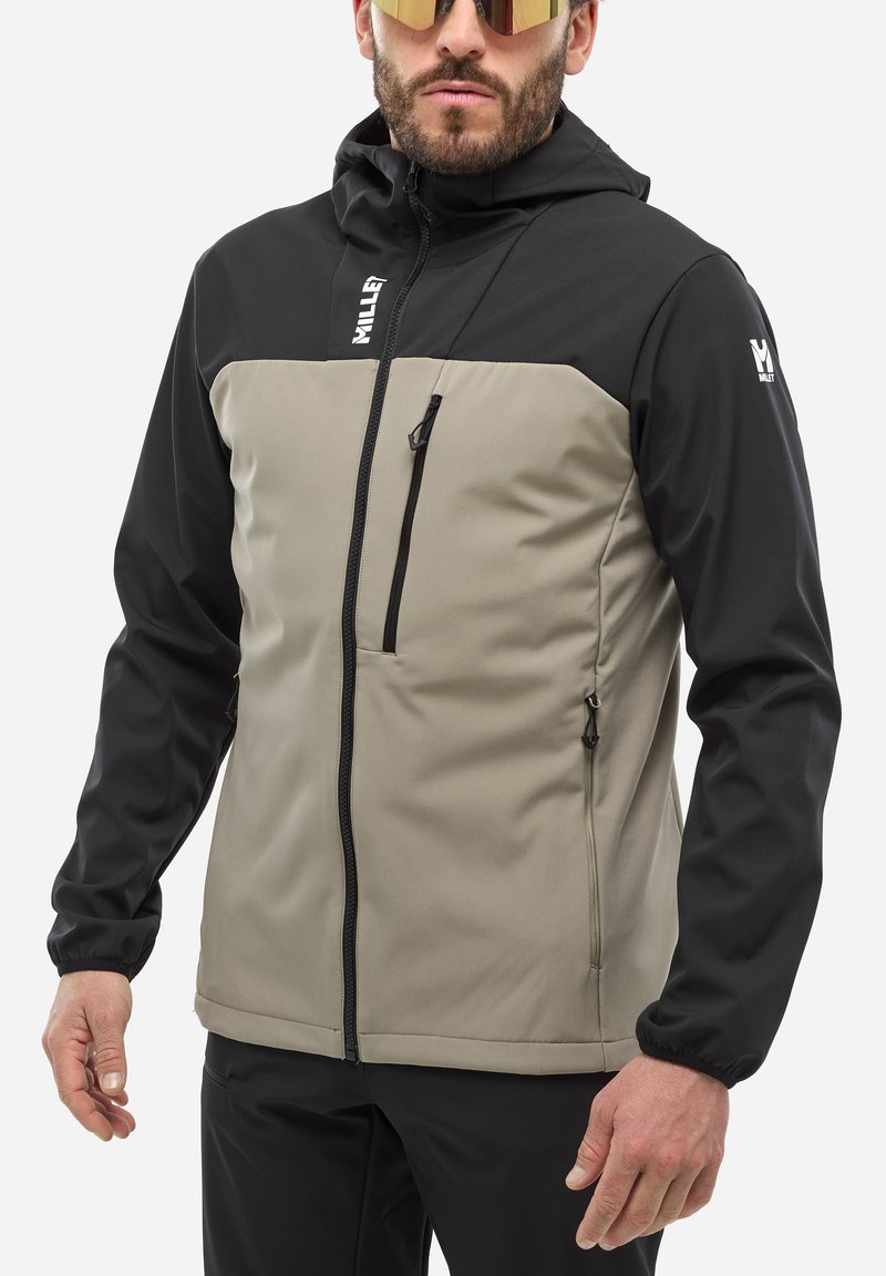 Veste à capuche noire et beige, en matériau softshell. Elle possède une poche poitrine zippée, des poignets réglables et un ourlet élastique.