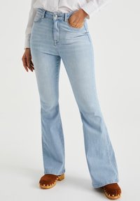 Light blue flared jeans with a high waistband, featuring front pockets and a subtle wash pattern. Paired with brown clogs.