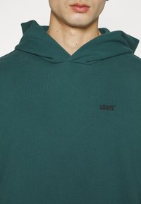 Kapturowa bluza w ciemnym odcieniu teal z teksturowanej tkaniny. Posiada małe wyszywane logo Levi's na klatce piersiowej. Klasyczny krój typu pullover.